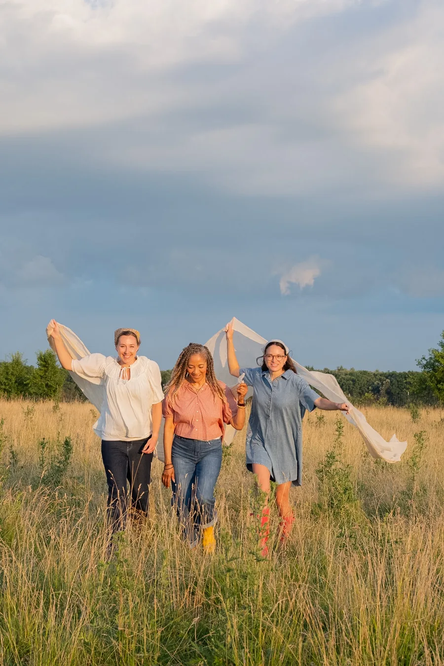 Three women lifting a flowing cloth together in a sunlit field for the 2027 photoshoot
